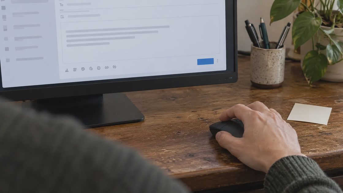 Review Before You Send A hand resting on a mouse beside a monitor displaying an indistinct email draft.