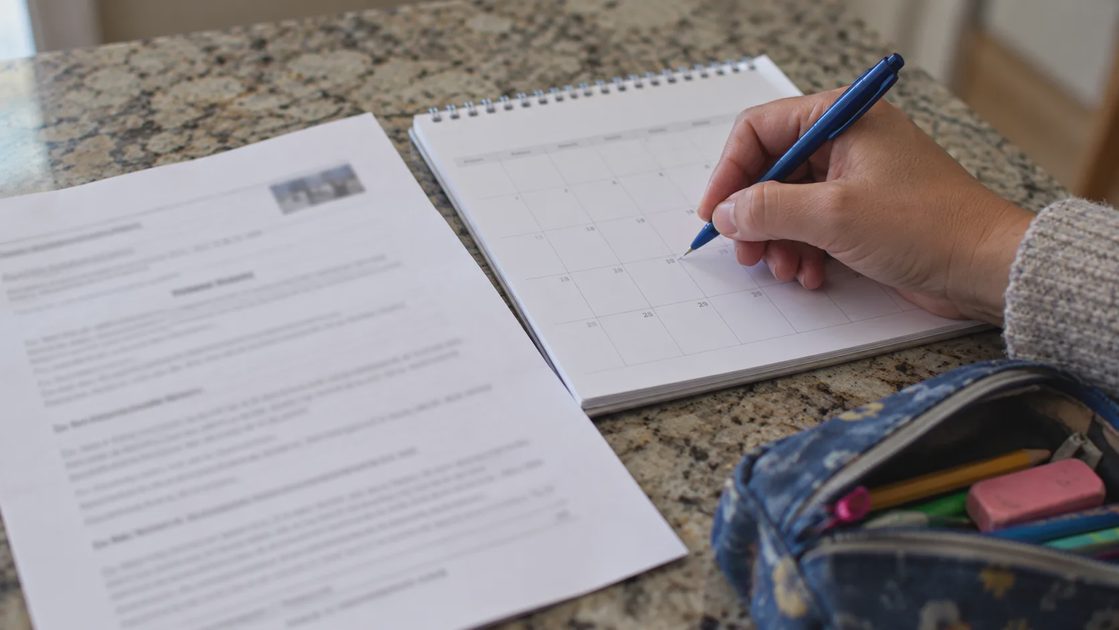 Verify the Deadline Before Relying on It A hand holding a pen checks a paper calendar beside a printed notice and pencil case.